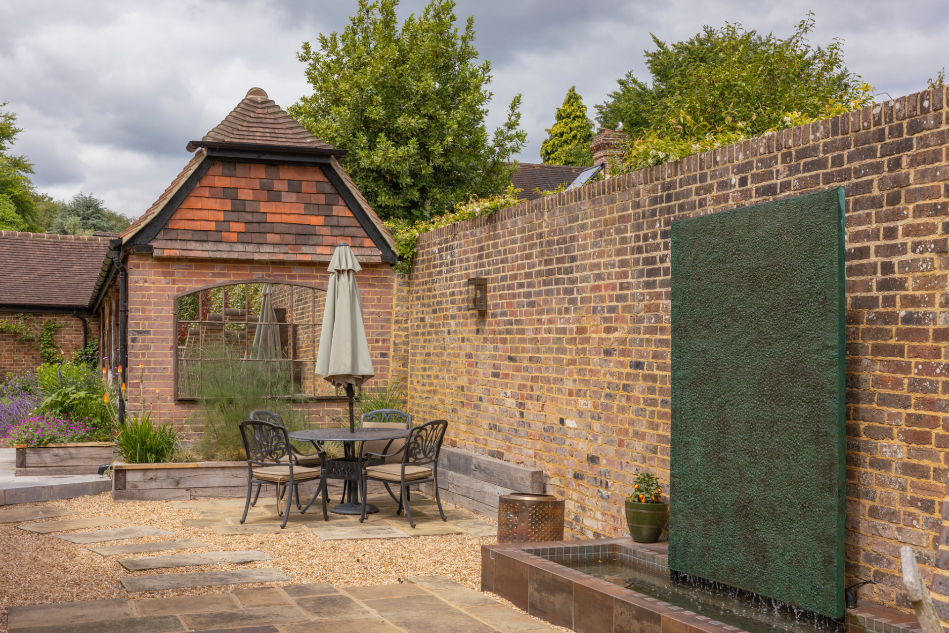 The finished sunlit brick courtyard built from that design, with the green metal water wall, raised pool, dining table, parasol and established planting