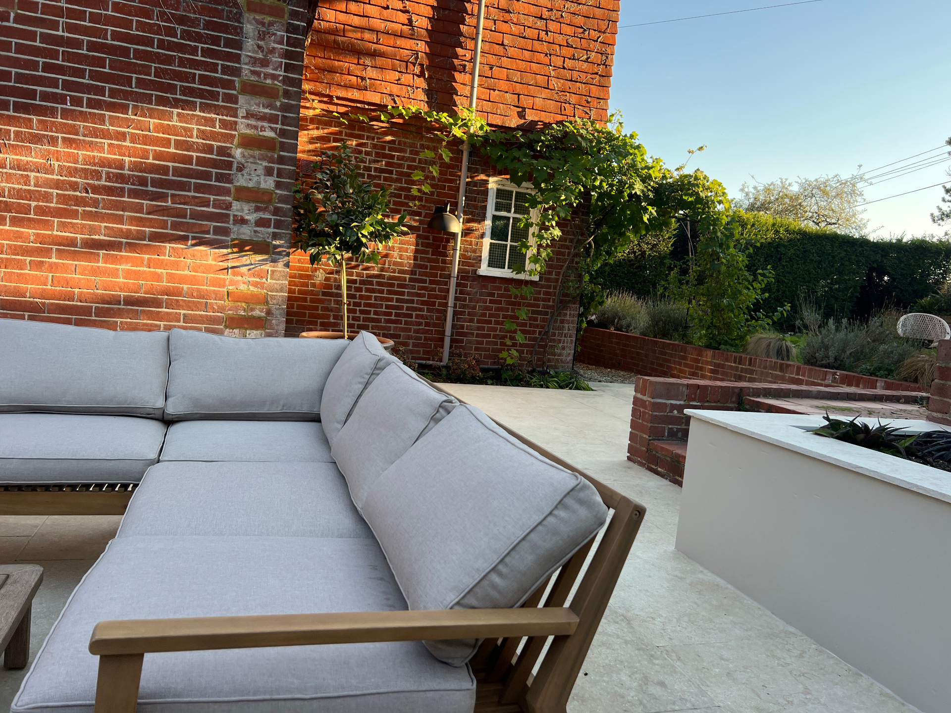 Medi Garden - travertine terrace with a low grey sofa beneath a vine-clad brick wall at golden hour